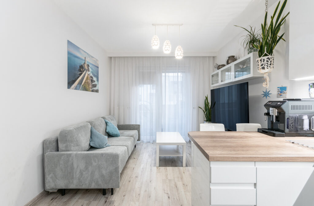 Jasny salon z szarą sofą, dużymi oknami i zasłonami.