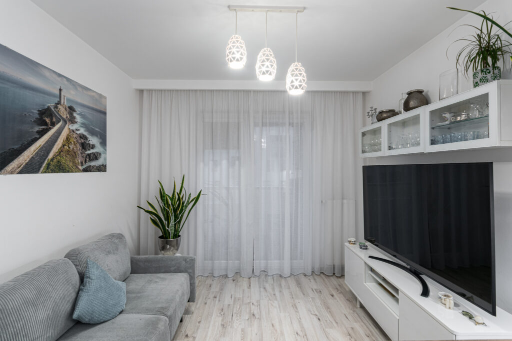 Salon z szarą sofą, roślinami doniczkowymi i przeszklonym wyjściem na balkon.