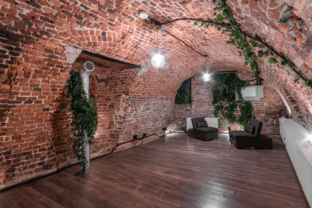 Ceglany, sklepiony salon w kamienicy z fotelami i zielonymi roślinami.